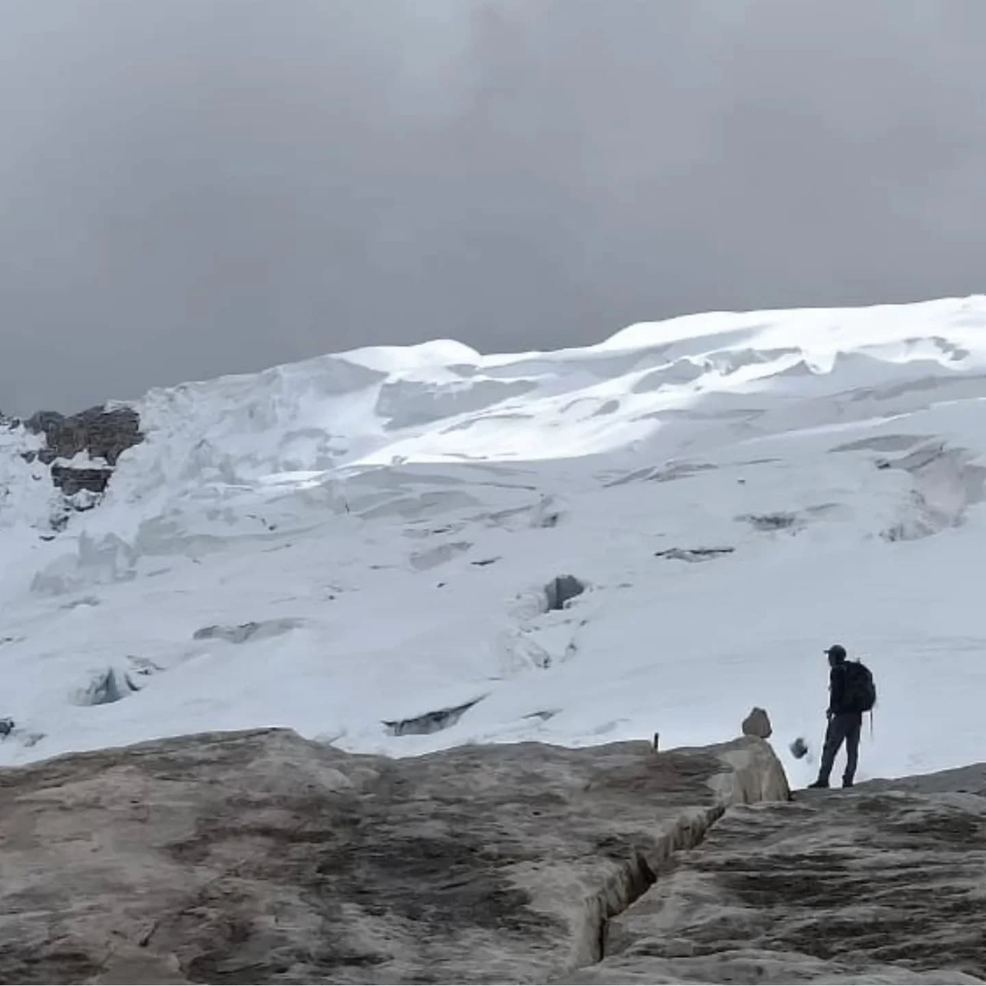 Campo de nieve y lengua de glaciar con una o dos personas en la base bajo cielo nublado.